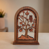 A small wooden decorative table piece featuring a laser-cut tree with autumn leaves and an owl inside an arched frame, displayed on a light tabletop with a softly blurred indoor background.