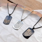 Three dog tag necklaces displayed on a white textured tray with a wooden surface in the background. The necklaces feature black, silver, and black metal tags with engraved American flag and eagle designs.