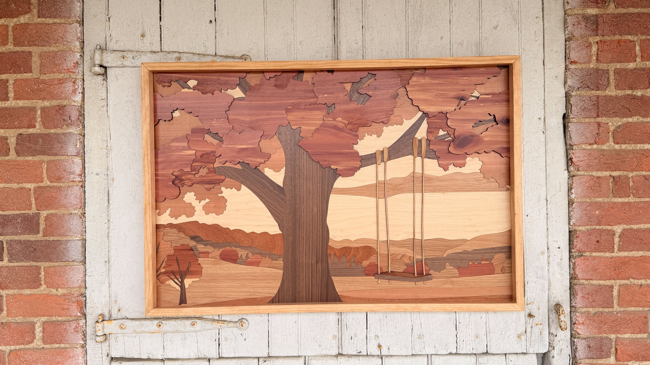 A rectangular wooden wall art piece featuring an inlaid landscape scene with a large tree and a swing, mounted on a white wooden door between red brick walls.