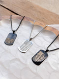 Three dog tag necklaces displayed on a white textured tray with a wooden surface in the background. The necklaces feature black, silver, and black metal tags with engraved American flag and eagle designs.